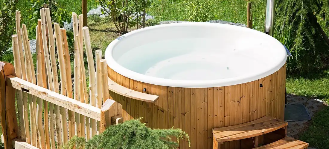 Outdoor wooden hot tub with steps on a deck, surrounded by green shrubs and trees.