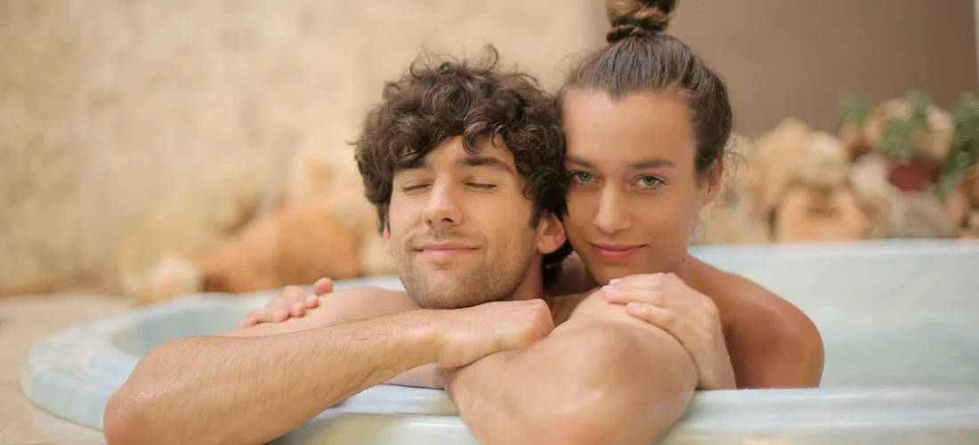 A man and a woman lean on the edge of a hot tub, smiling with their arms resting on the rim.