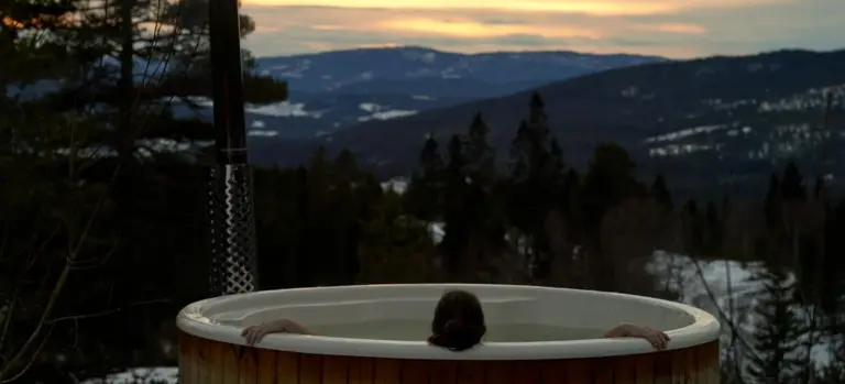 A person sits in an outdoor wooden hot tub at dusk, with pine trees and distant mountains in the background.