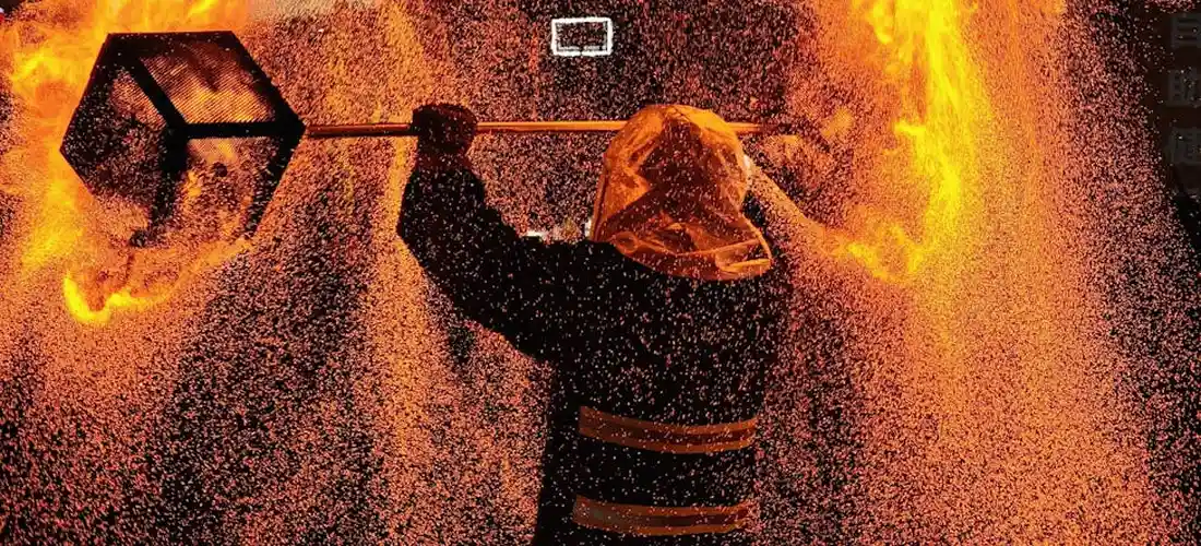Firefighter in turnout gear and protective hood fighting a blaze with a long tool, surrounded by intense flames