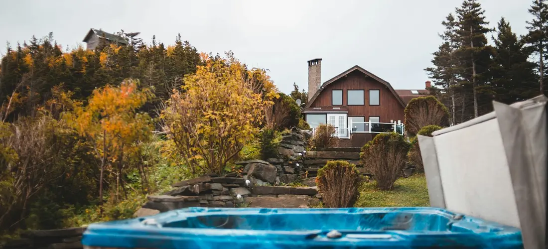 Backyard scene with a blue hot tub in the foreground, autumn trees, and a house in the background.