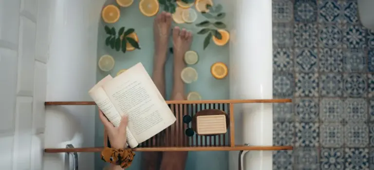 Top-down view of a person soaking in a hot tub with a wooden tray across the tub holding a book; lemon slices float on the water; blue patterned tiles surround the tub.