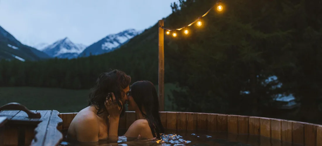 Two adults soaking in an outdoor hot tub at dusk with string lights and mountain scenery in the background.