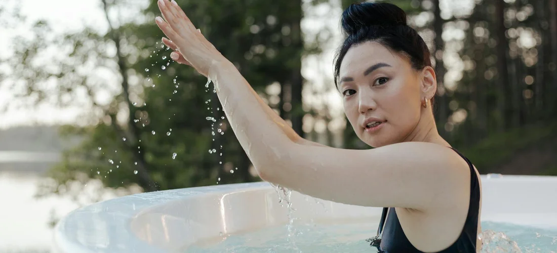 Woman in an outdoor hot tub wearing a dark swimsuit, raising her hand as water splashes; trees and water in the background.