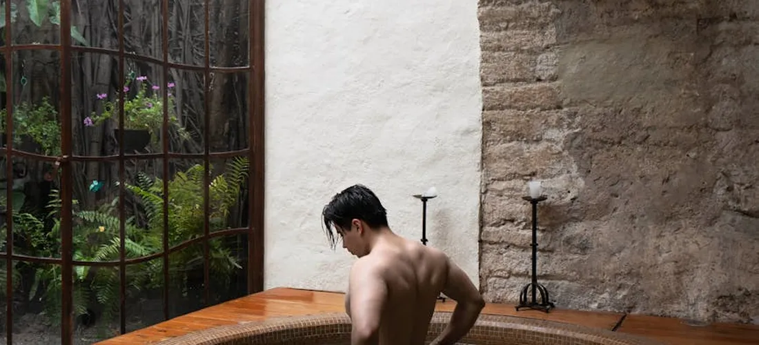 Man soaking in a wooden hot tub indoors, back turned, next to a window with a stone wall exterior.
