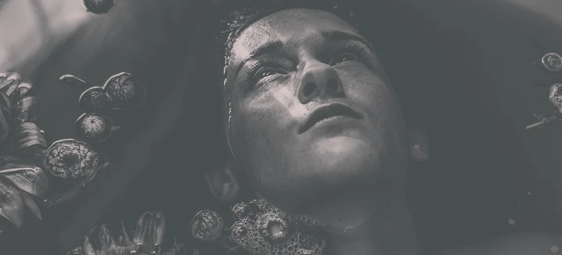 A person’s face partially submerged in water, surrounded by bubbles and dark organic shapes, shown in grayscale.