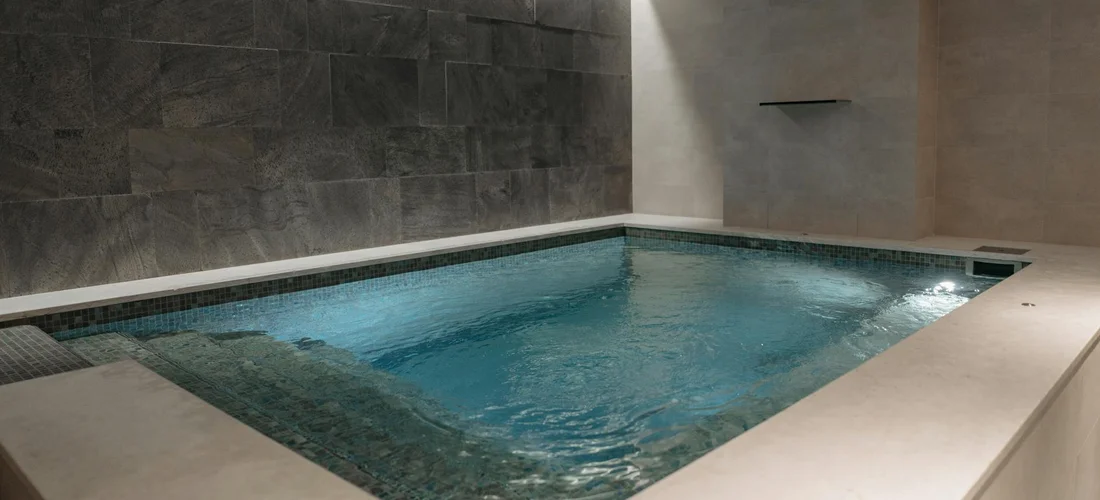 Indoor hot tub with clear blue water, surrounded by light concrete edges and dark tiled wall.