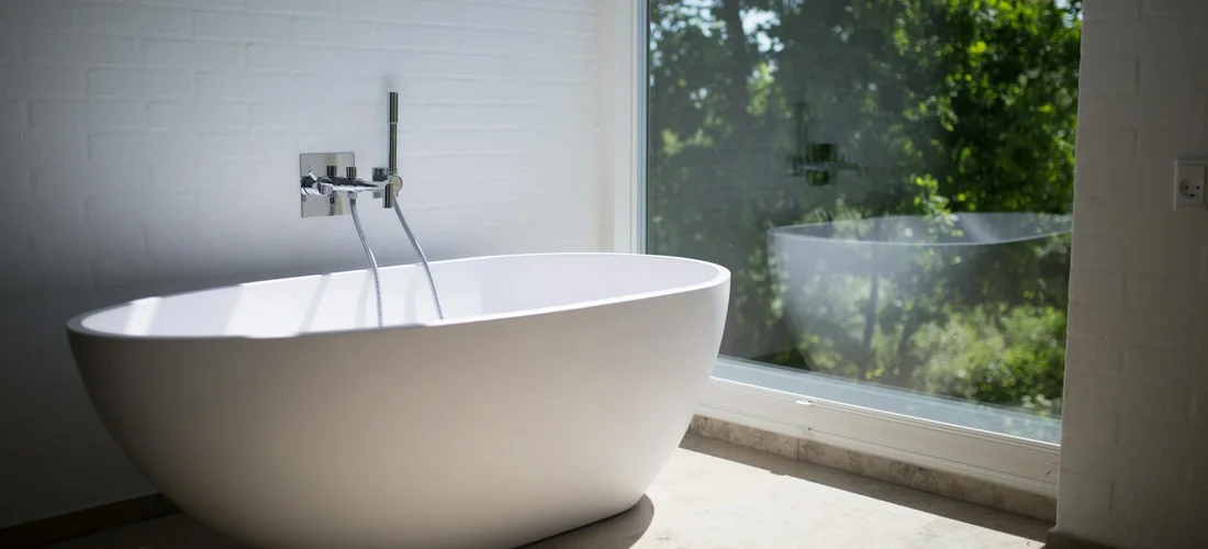 White freestanding bathtub in a bright bathroom next to a large window overlooking greenery.