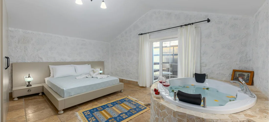 A bedroom with a round Jacuzzi tub on the right, a large bed on the left, and a window with sheer curtains, featuring patterned wallpaper and tiled flooring.