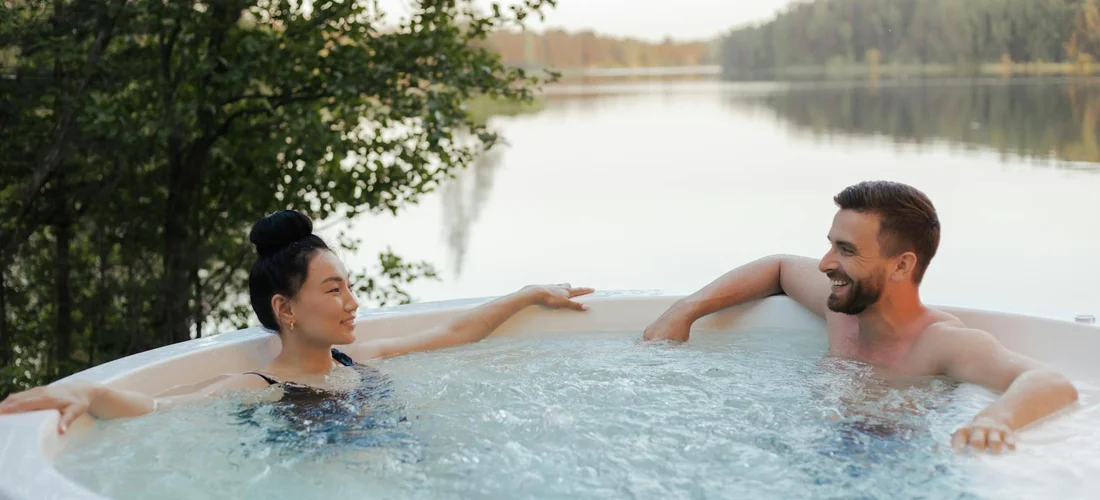 Two adults relax in an outdoor Jacuzzi on a sunny day, with a lake and trees in the background.