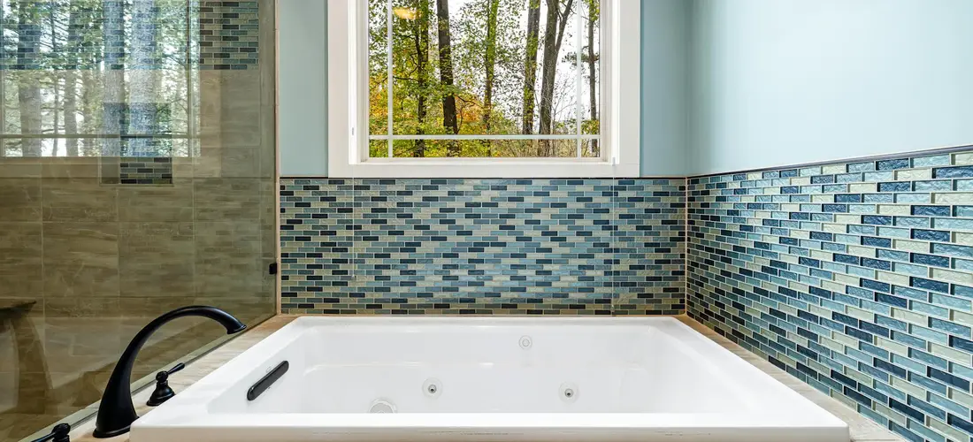 A white Jacuzzi tub surrounded by blue mosaic tile in a bright bathroom with a window above.