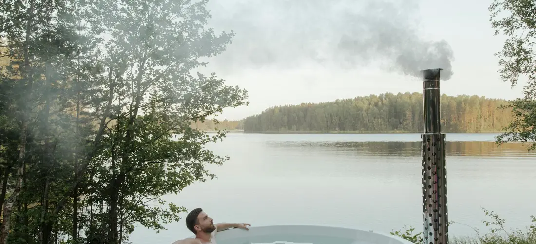 Outdoors hot tub by a calm lake with steam rising from the water; a person sits in the tub, relaxing as mist fills the air.