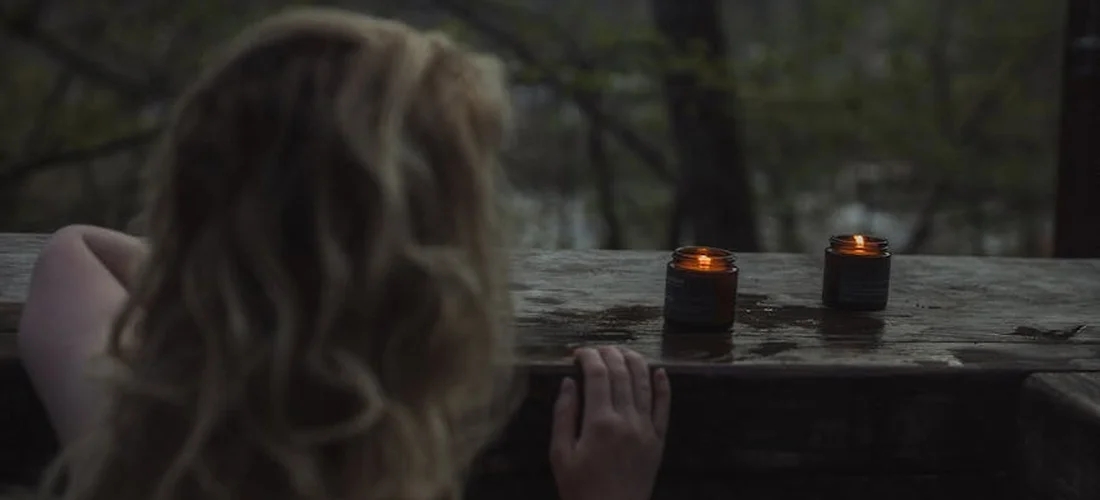 Back view of a person with light hair looking out over a wooden railing at dusk, with small candles on the railing.