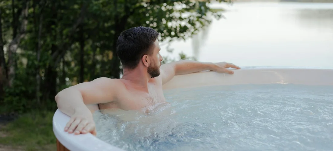 Man relaxing in an outdoor hot tub with arms resting on the rim, water rippling, trees and a lake in the background.