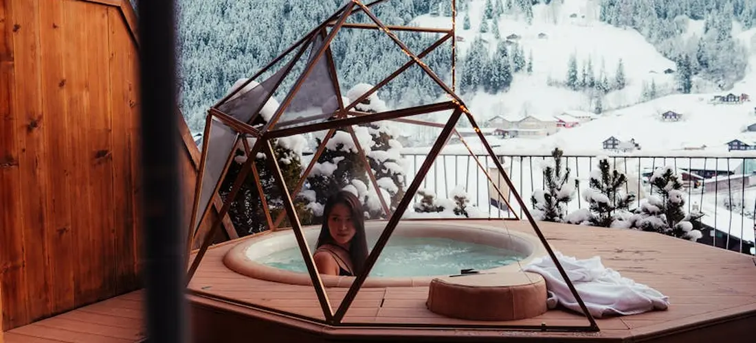 Person relaxing in a hot tub on a wooden deck with a geometric metal-frame lounge structure and snowy mountains in the background.
