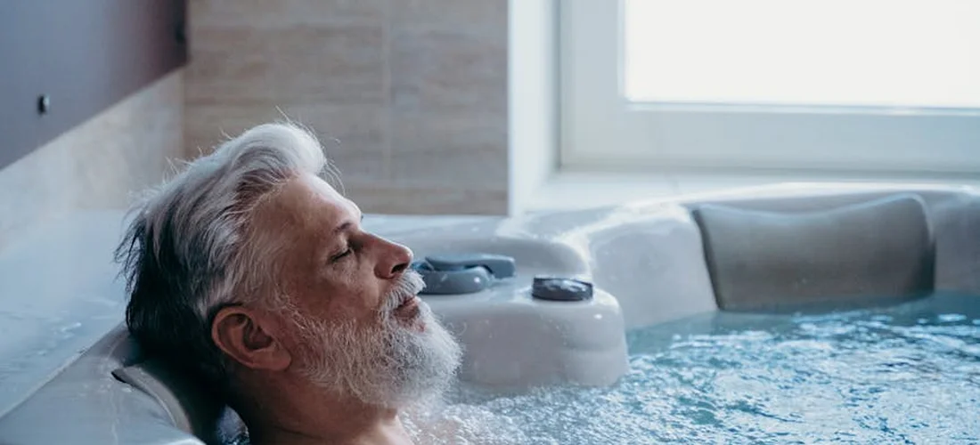 An older man with a white beard relaxes in a Jacuzzi, looking toward a bright window.