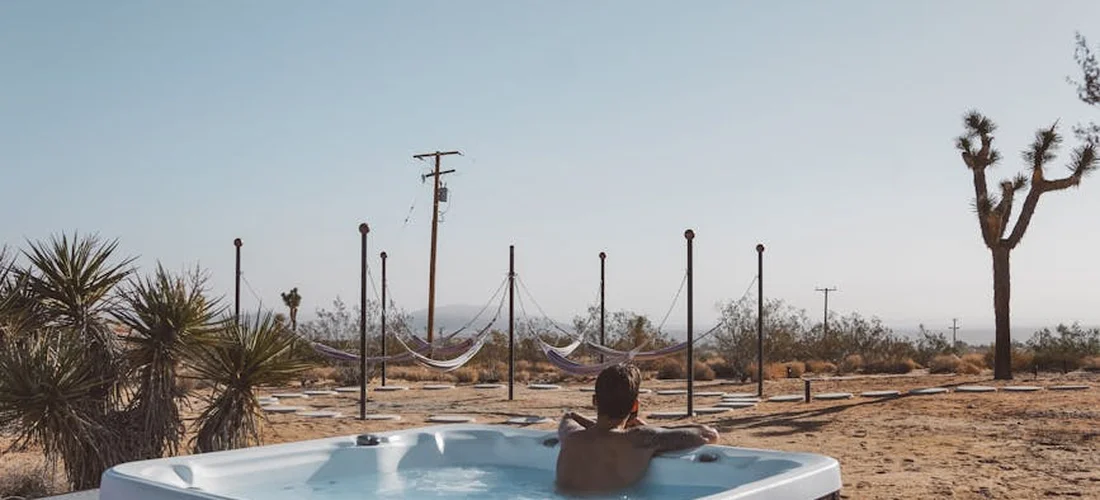 A person sits in an outdoor hot tub in a desert landscape with palm trees and a clear blue sky.