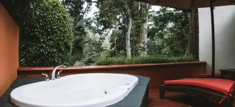 Outdoor jacuzzi tub on a wooden deck with lush greenery in the background and a red lounge chair nearby.