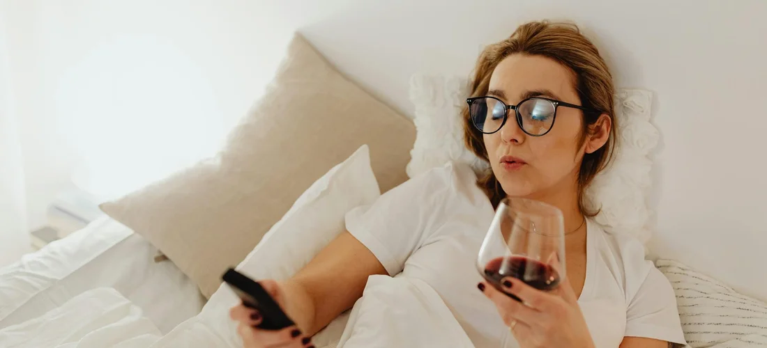 Woman wearing glasses sits on a bed, holding a TV-style remote in one hand and a wine glass in the other.