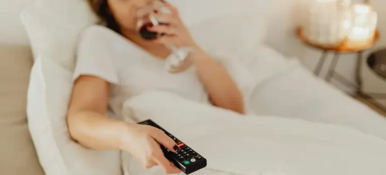Close-up of a hand holding a remote control in the foreground with a person relaxing on a couch in the blurred background.