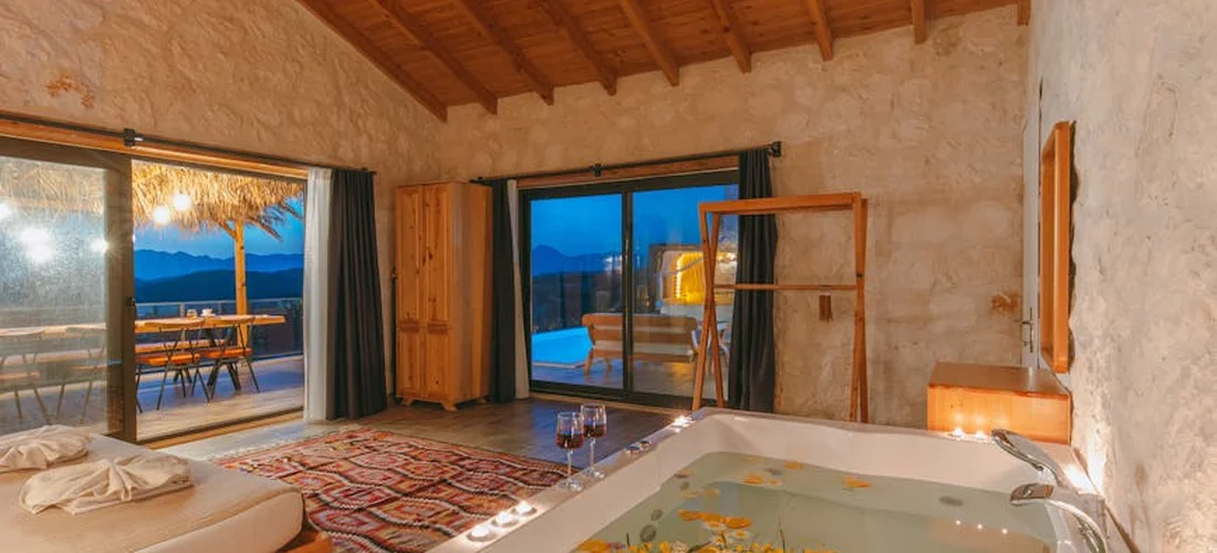 A luxury Jacuzzi suite with a large built-in tub, stone walls, wooden ceiling beams, and glass doors opening to a balcony with a mountain view.