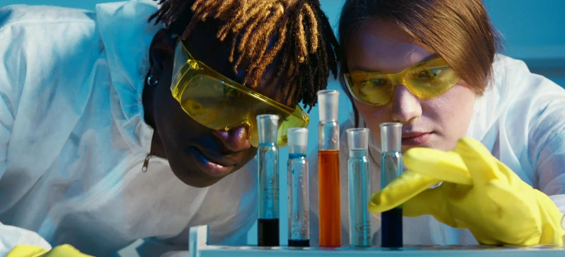 Two researchers wearing safety goggles and yellow gloves examine test tubes containing colorful liquids in a laboratory.