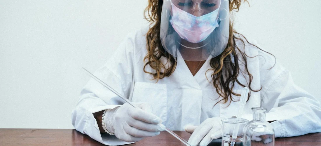 A lab technician wearing a white coat, gloves, and a face shield uses a pipette at a lab bench with glassware nearby.