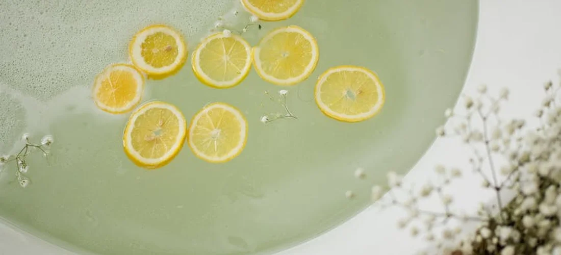 Top-down view of lemon slices floating in a round glass dish with a pale green liquid, with a blurred plant in the background.