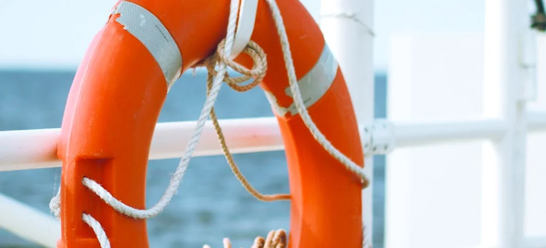 Orange lifebuoy with white rope hanging on a railing with water in the background.