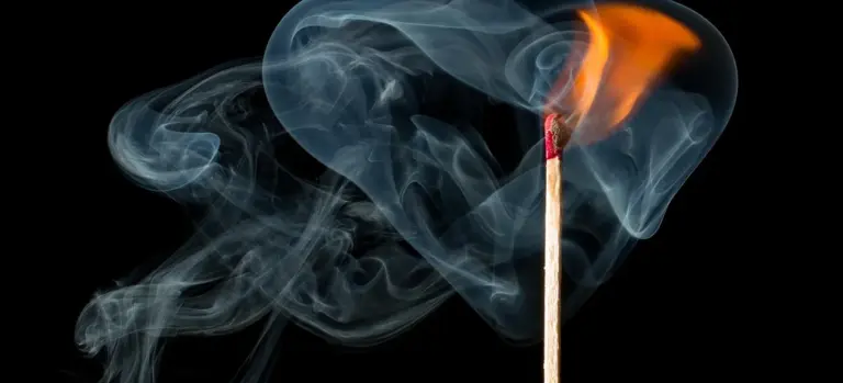 A lit wooden match with orange flame and blue smoke rising against a black background.