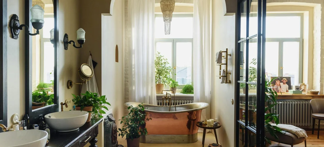 Luxurious home bathroom with a freestanding oval tub centered between large windows, flanked by potted plants and a double vanity, bathed in natural light.