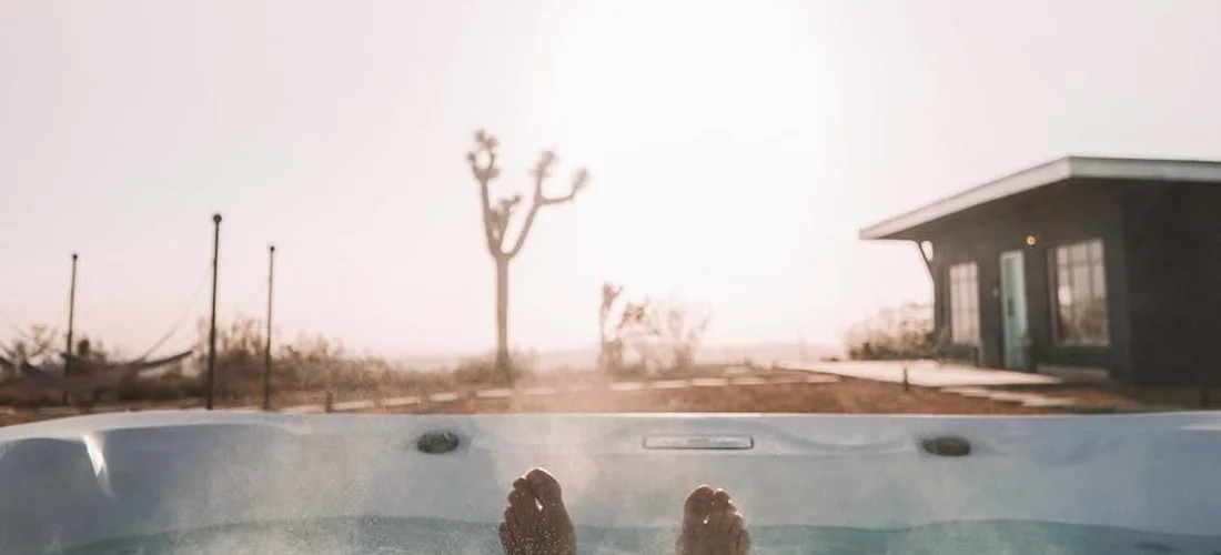 Feet resting in an outdoor hot tub, looking toward a sunny yard with a small building in the distance.