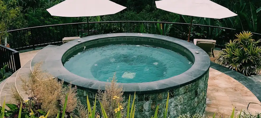 Circular outdoor hot tub with a stone surround set in a garden, with patio umbrellas and lush plants nearby.