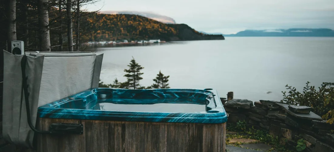 Blue hot tub installed outdoors by a scenic lake with trees and cliffside, illustrating the importance of reliable service and maintenance for hot tubs.