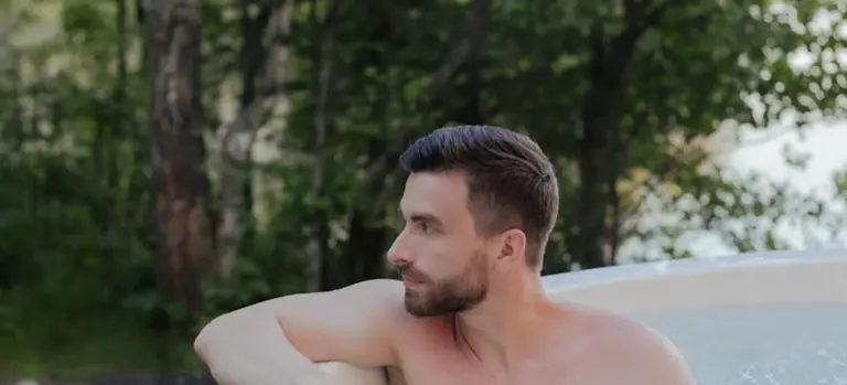 Adult man relaxing in an outdoor hot tub surrounded by trees and greenery.