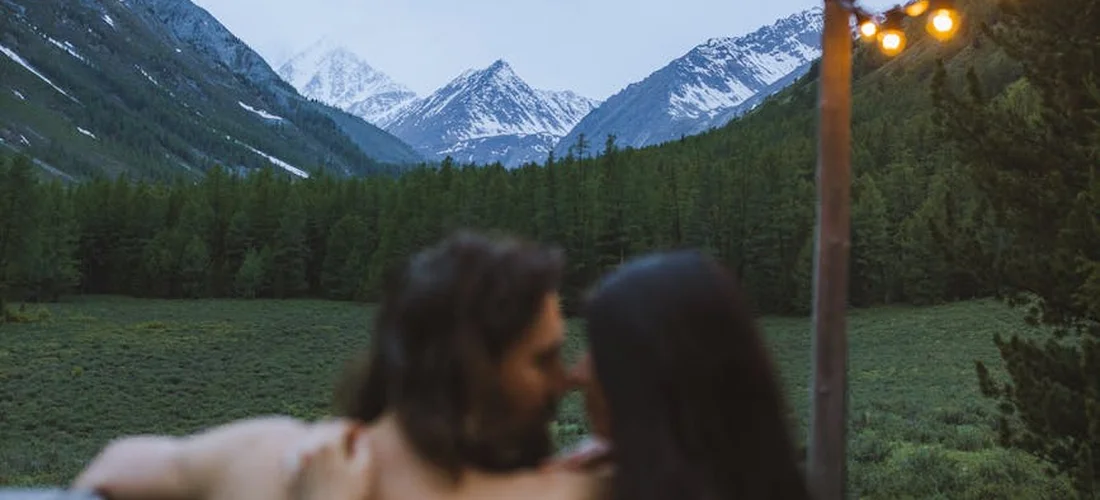 Blurry foreground of two people in an outdoor hot tub, with a scenic mountain landscape and evergreen trees in the background.
