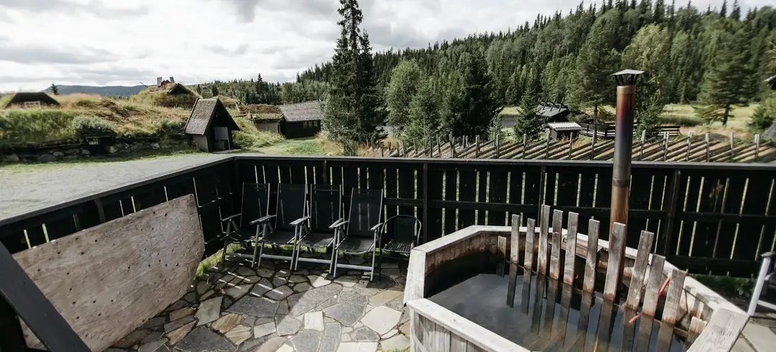 Outdoor wooden hot tub on a stone patio with a dark wooden fence and a forested hillside in the background.