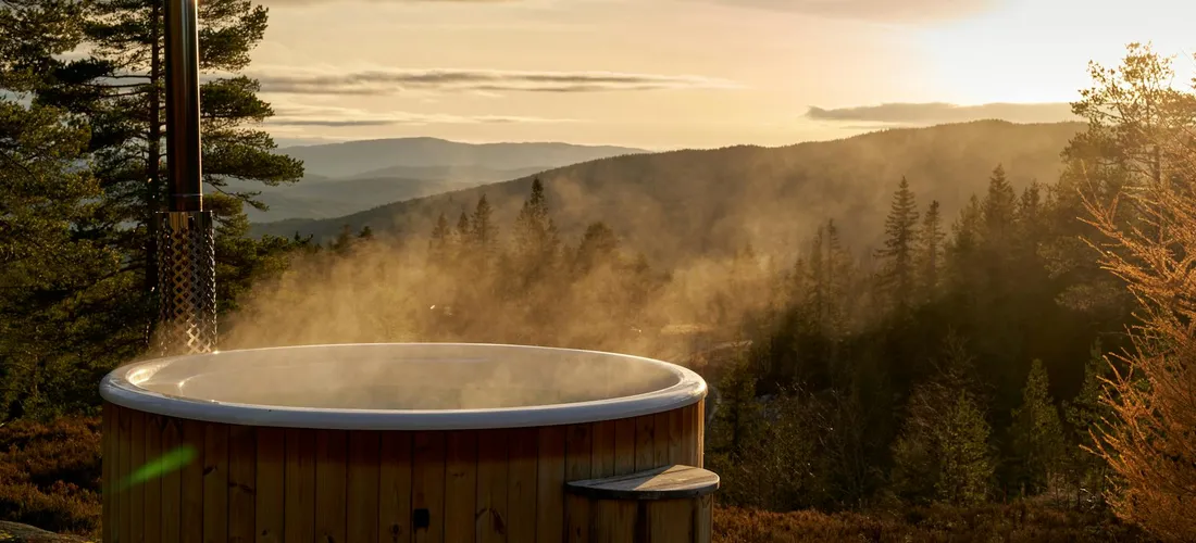 A wooden outdoor hot tub releasing steam, set on a deck with a forested mountain landscape and a warm sunset.
