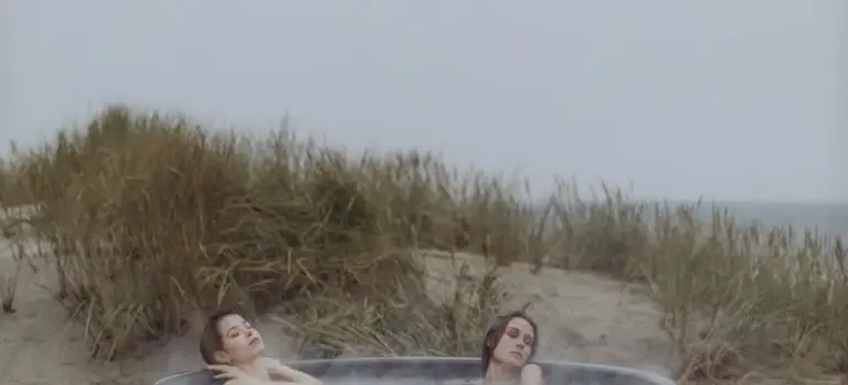 Two people soak in an outdoor hot tub with dune grasses and a cloudy sky in the background.
