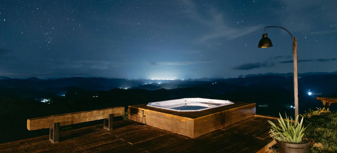 Nighttime outdoor hot tub on a wooden deck with a bench, lamp, and potted plant under a star-filled sky.