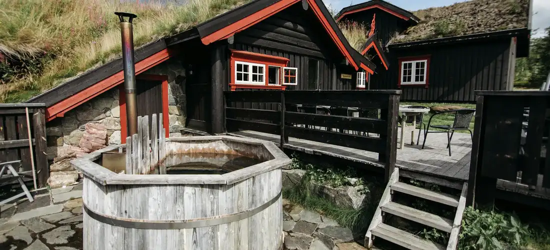 Outdoor wooden barrel hot tub with a metal stove on a stone patio, adjacent wooden deck stairs, and cabin-style buildings in the background.
