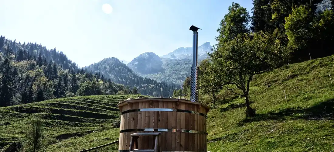 Outdoor wooden hot tub on a grassy hillside with trees and distant mountains
