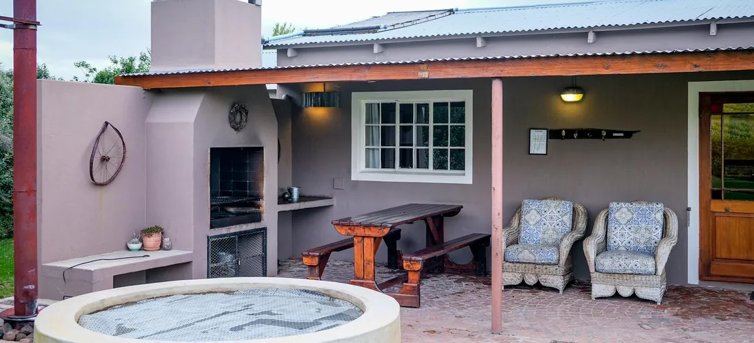 Backyard patio with a circular hot tub in the foreground, a built-in grill on the left, a wooden picnic table under a covered seating area, and two cushioned chairs.