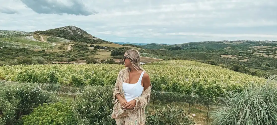 Woman standing in a sunlit vineyard landscape with rolling hills, wearing a white sleeveless top and a light beige cardigan.