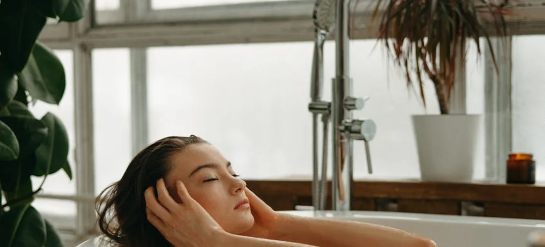 A person relaxing in a bathtub in a bright bathroom with natural light, modern fixtures, and potted plants.