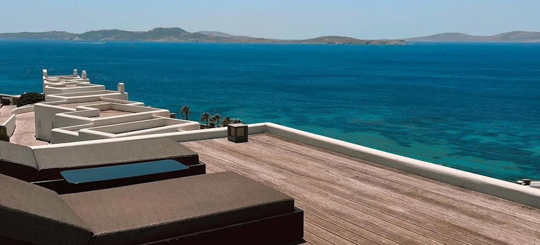 A wooden rooftop deck overlooking a calm blue sea with distant islands on the horizon and lounge seating along the railing.