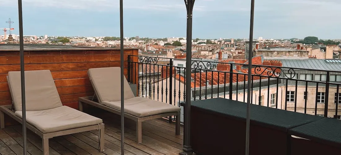 Rooftop deck with wooden planks, two lounge chairs, a metal railing, and an urban cityscape in the distance.