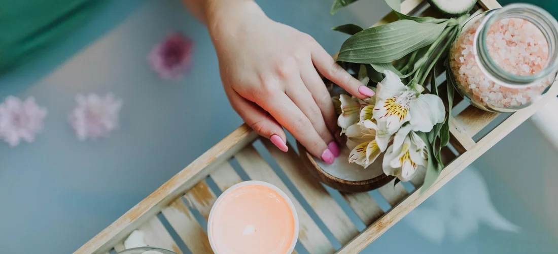 Close-up of a hand with pink manicure resting on a wooden bath tray adorned with flowers and jars of bath products beside a tub.