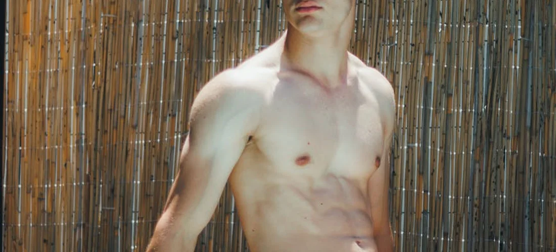 A shirtless man standing outdoors in front of a vertical wooden fence