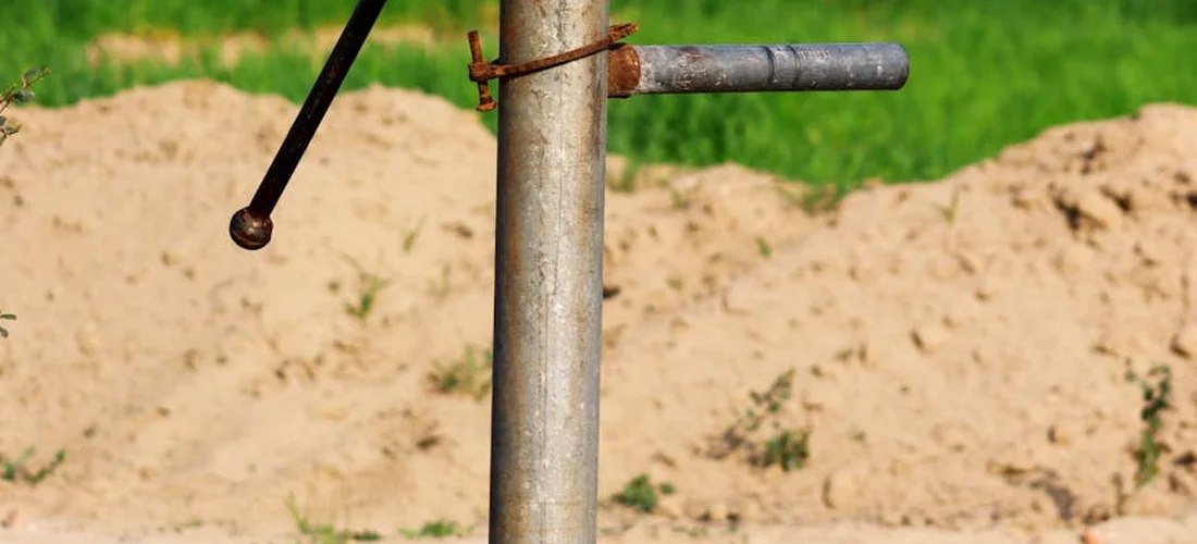 Close-up of a manual hand pump lever attached to a vertical metal pipe with a sandy background.